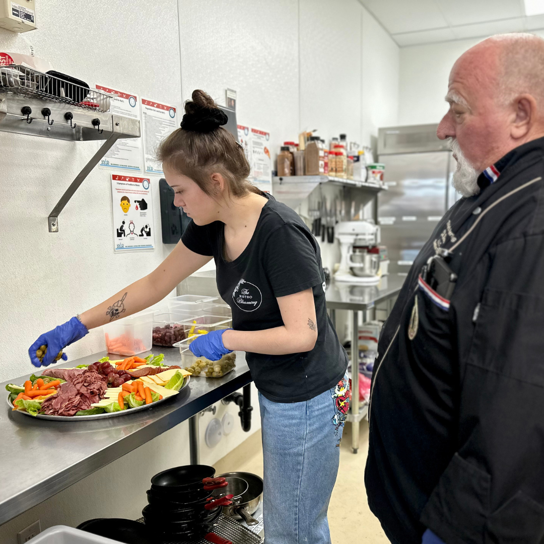 A male chef teaches a female employee how to make a charcuterie board